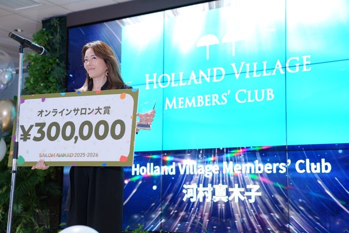 DMMオンラインサロン「SALON AWARD 2025-2026」の授賞式ステージで表彰される河村真木子氏。背景に「HOLLAND VILLAGE MEMBERS' CLUB」の文字